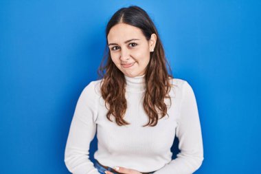 Young hispanic woman standing over blue background with hand on stomach because indigestion, painful illness feeling unwell. ache concept. 