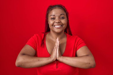 African american woman with braided hair standing over red background praying with hands together asking for forgiveness smiling confident. 