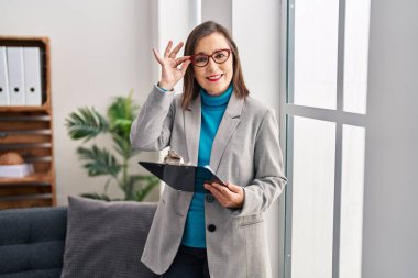 Middle age woman psychologist smiling confident holding clipboard at psychology center
