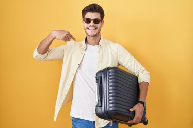 Young hispanic man holding suitcase going on summer vacation looking confident with smile on face, pointing oneself with fingers proud and happy. 