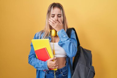 Young blonde woman wearing student backpack and holding books bored yawning tired covering mouth with hand. restless and sleepiness. 