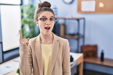 Teenager girl working at the office wearing glasses pointing finger up with successful idea. exited and happy. number one. 