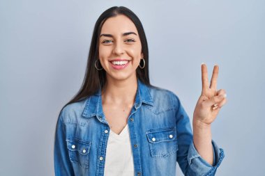 Mavi arka planda duran İspanyol kadın iki numarayı gösterip işaret ederken kendinden emin ve mutlu bir şekilde gülümsüyor.. 