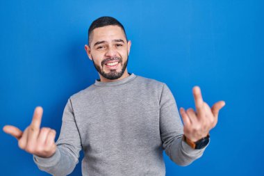 Hispanic man standing over blue background showing middle finger doing fuck you bad expression, provocation and rude attitude. screaming excited 