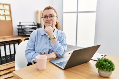 Young redhead woman working at the office using computer laptop looking confident at the camera smiling with crossed arms and hand raised on chin. thinking positive. 