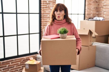 Middle age hispanic woman moving to a new home holding cardboard box making fish face with mouth and squinting eyes, crazy and comical. 