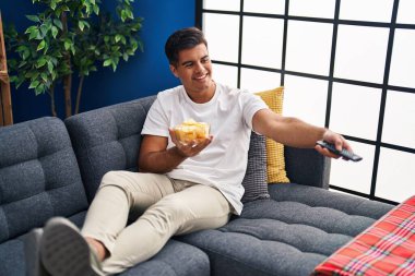 Young hispanic man watching movie eating chips potatoes at home