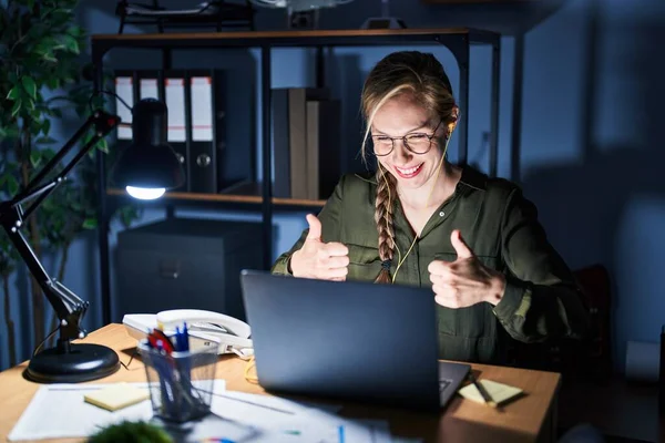 Young blonde woman working at the office at night approving doing positive gesture with hand, thumbs up smiling and happy for success. winner gesture.