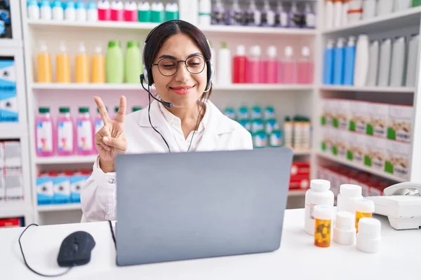 Young arab woman working at pharmacy drugstore using laptop showing and pointing up with fingers number two while smiling confident and happy.