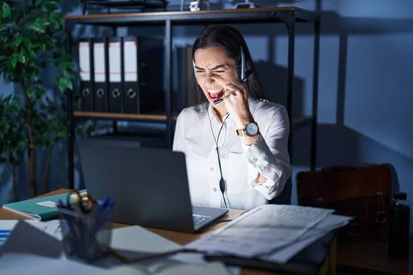 Young brunette woman wearing call center agent headset working late at night angry and mad raising fist frustrated and furious while shouting with anger. rage and aggressive concept.