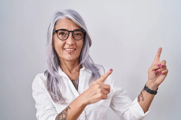 Middle age woman with tattoos wearing glasses standing over white background smiling and looking at the camera pointing with two hands and fingers to the side.