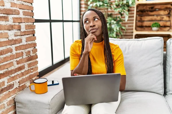 非洲妇女坐在沙发上 在家里用笔记本电脑 严肃地面面面相觑 手托着下巴思考着问题 思考着困惑的想法 — 图库照片