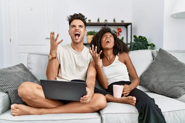 stock image Young interracial couple using laptop at home sitting on the sofa crazy and mad shouting and yelling with aggressive expression and arms raised. frustration concept. 