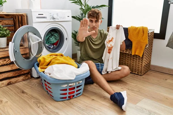 Young caucasian man doing laundry holding t shirt with stain with open hand doing stop sign with serious and confident expression, defense gesture