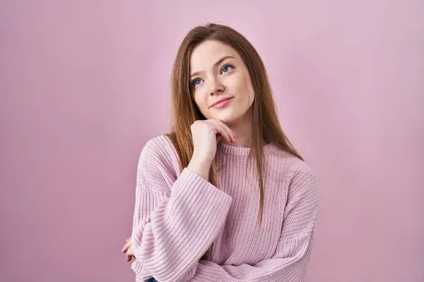 若い白人女性はピンクの背景の上に立っていて 顎は質問 集中的な表現について考えています 思慮深い顔で笑顔 疑わしい概念 — ストック写真