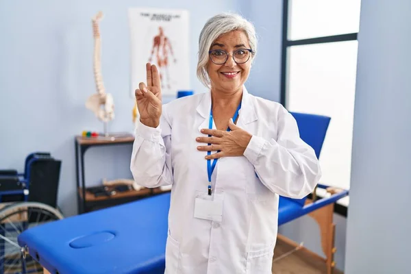 Middle age woman with grey hair working at pain recovery clinic smiling swearing with hand on chest and fingers up, making a loyalty promise oath