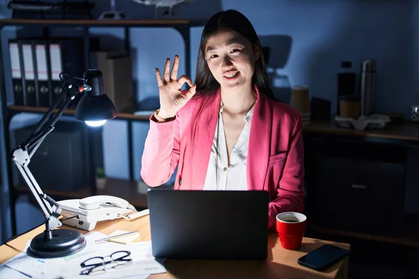 Chinese young woman working at the office at night smiling positive doing ok sign with hand and fingers. successful expression.