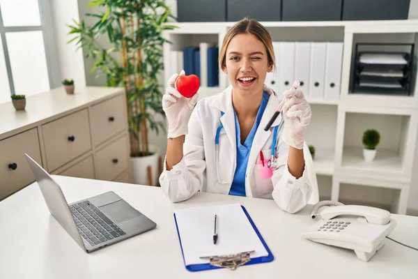 Young Blonde Woman Wearing Doctor Uniform Holding Blood Heart Winking — Stock Photo, Image