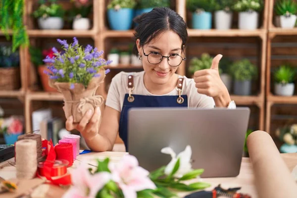 Young hispanic woman working at florist shop doing video call approving doing positive gesture with hand, thumbs up smiling and happy for success. winner gesture.