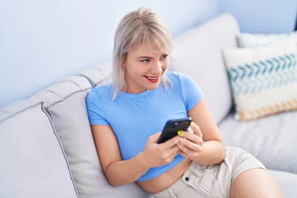 年轻的金发女人坐在家里的沙发上用智能手机 — 图库照片