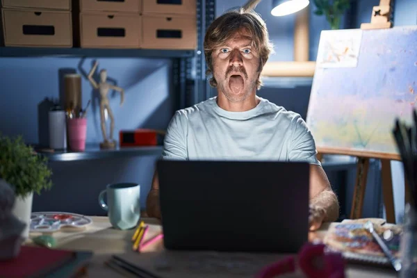 深夜にノートパソコンを持ってアートスタジオに座っている中年男性が面白い表情で幸せ舌を突き出しています 感情の概念 — ストック写真