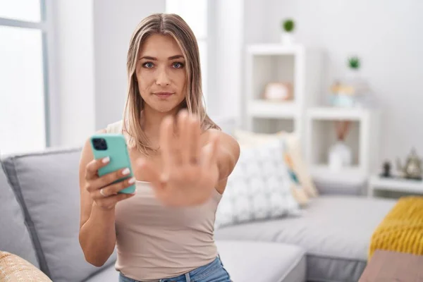 Blonde caucasian woman using smartphone typing message sitting on the sofa with open hand doing stop sign with serious and confident expression, defense gesture
