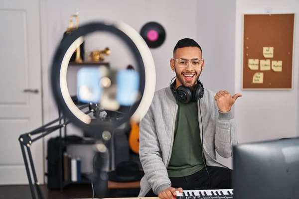 Young hispanic man playing piano at music studio recording himself pointing thumb up to the side smiling happy with open mouth