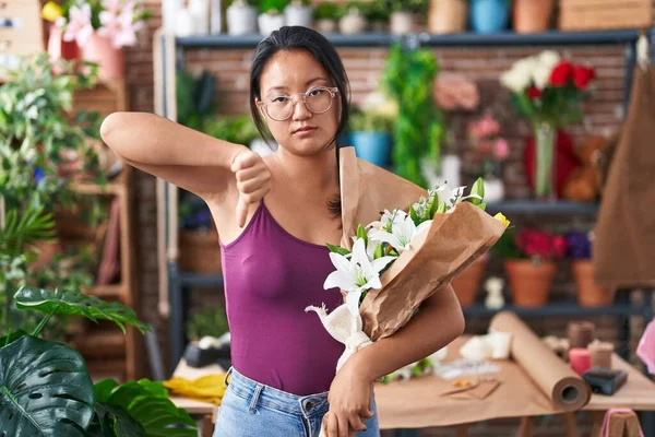 Asian young woman at florist shop holding bouquet of flowers with angry face, negative sign showing dislike with thumbs down, rejection concept