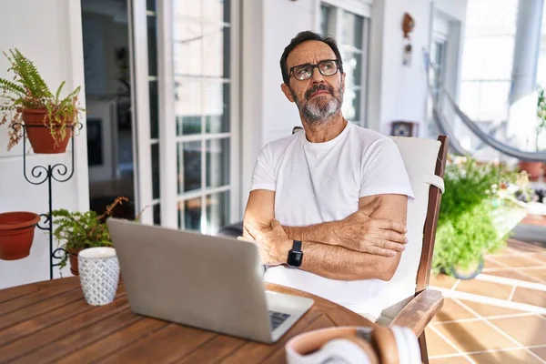 Middle age man using computer laptop at home looking to the side with arms crossed convinced and confident