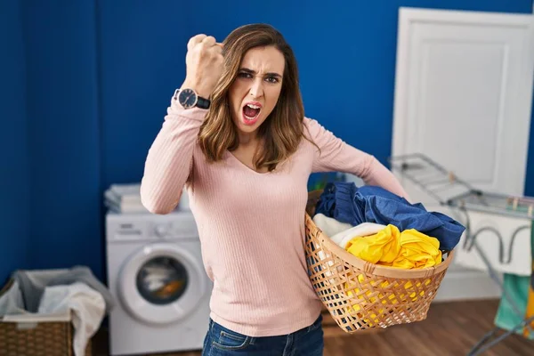 Young woman holding laundry basket angry and mad raising fist frustrated and furious while shouting with anger. rage and aggressive concept.