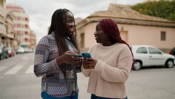 İki Afrikalı Amerikalı arkadaş sokakta akıllı telefon kullanarak gülümsüyor.