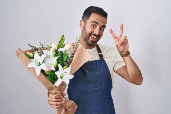 Hispanic man with beard working as florist smiling looking to the camera showing fingers doing victory sign. number two.