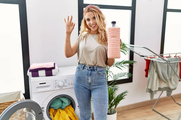 Beautiful blonde woman doing laundry holding detergent bottle doing ok sign with fingers, smiling friendly gesturing excellent symbol