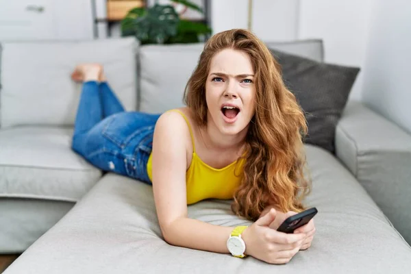 Young caucasian woman lying on the sofa using smartphone angry and mad screaming frustrated and furious, shouting with anger. rage and aggressive concept.