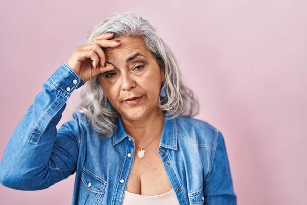 Middle age woman with grey hair standing over pink background worried and stressed about a problem with hand on forehead, nervous and anxious for crisis