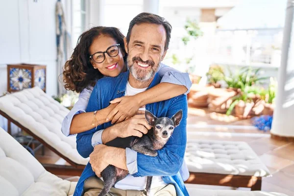 Middle age hispanic couple hugging each other sitting on hammock with dog at terrace