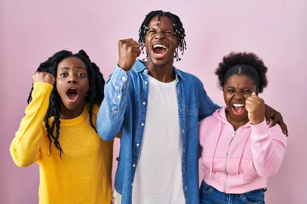 Group of three young black people standing together over pink background angry and mad raising fist frustrated and furious while shouting with anger. rage and aggressive concept.