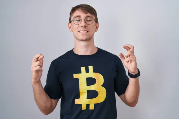 stock image Caucasian blond man wearing bitcoin t shirt gesturing finger crossed smiling with hope and eyes closed. luck and superstitious concept. 