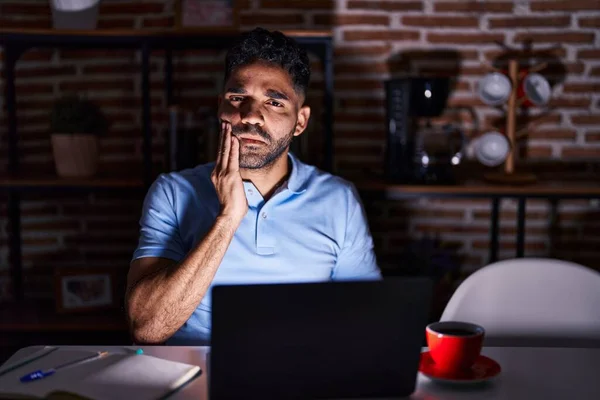 Hispanic man with beard using laptop at night touching mouth with hand with painful expression because of toothache or dental illness on teeth. dentist