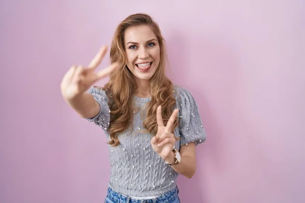 Beautiful blonde woman standing over pink background smiling with tongue out showing fingers of both hands doing victory sign. number two.