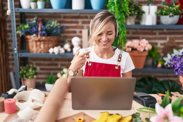 Young blonde woman working at florist shop doing video call smiling happy pointing with hand and finger