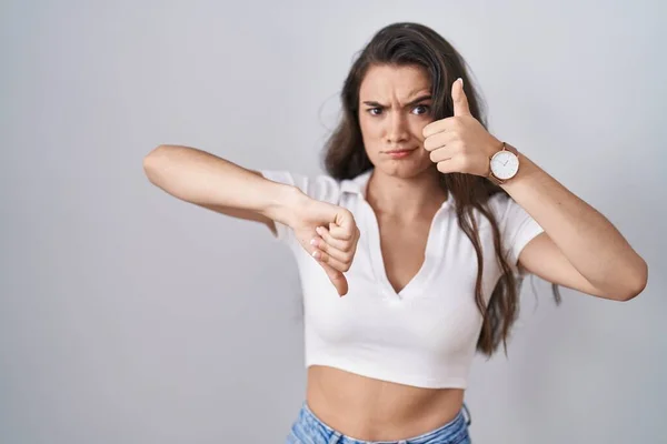 Young teenager girl standing over white background doing thumbs up and down, disagreement and agreement expression. crazy conflict