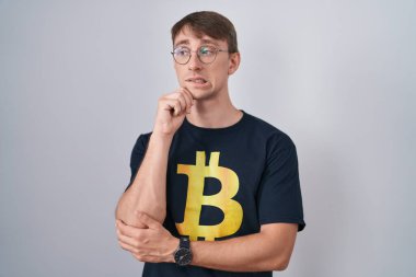 Caucasian blond man wearing bitcoin t shirt thinking worried about a question, concerned and nervous with hand on chin 