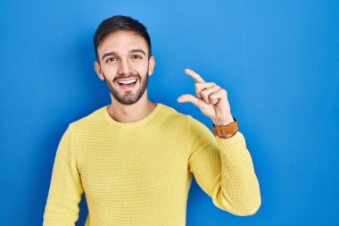 Hispanic man standing over blue background smiling and confident gesturing with hand doing small size sign with fingers looking and the camera. measure concept. 