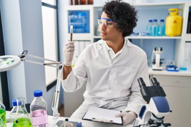 Laboratuvarda elinde pano ve test tüpü tutan bilim adamı üniforması giyen İspanyol genç adam.