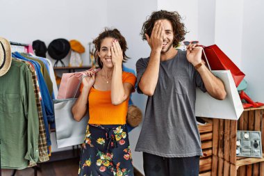 Genç İspanyol çift alışveriş torbalarını ellerinde tutarken bir gözünü eliyle kapatıyor, yüzünde kendine güvenen bir gülümseme ve sürpriz bir duygu oluşuyor.. 