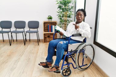 Young black woman sitting on wheelchair at waiting room surprised pointing with finger to the side, open mouth amazed expression. 