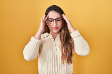 Young hispanic woman standing over yellow background with hand on head for pain in head because stress. suffering migraine. 