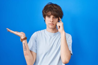 Mavi arka planda dikilen İspanyol genç adam avuç içinin açık olmasından rahatsız ve kafası karışmış bir şekilde fotokopi alanını gösteriyor ve alnını işaret ediyor. Bunu bir düşün.. 