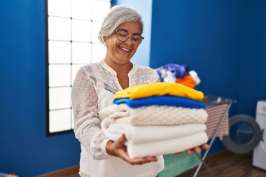 Çamaşırhanede katlanmış giysileri tutan orta yaşlı bir kadın gülümsüyor.